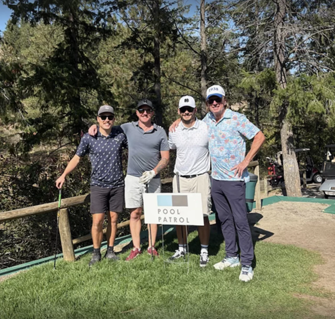 Four members of the Pool Patrol team stand arm in arm on a sunny tee box at St. Andrews by the Lake, smiling behind a Pool Patrol hole sponsor sign, with pine trees and golf carts in the background.