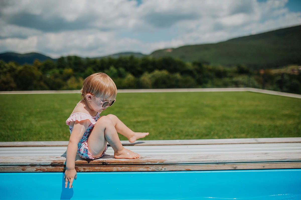 A young child in a swimsuit sitting at the edge of an open pool, feet near the water.