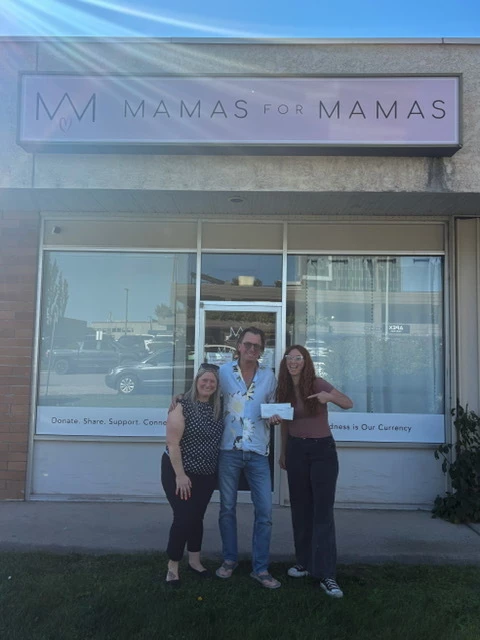 Allan Horwood, owner of Pool Patrol, stands outside the Mamas for Mamas storefront in Kelowna on a sunny day, holding a cheque between two members of the Mamas for Mamas team, one of whom smiles and points at the cheque.